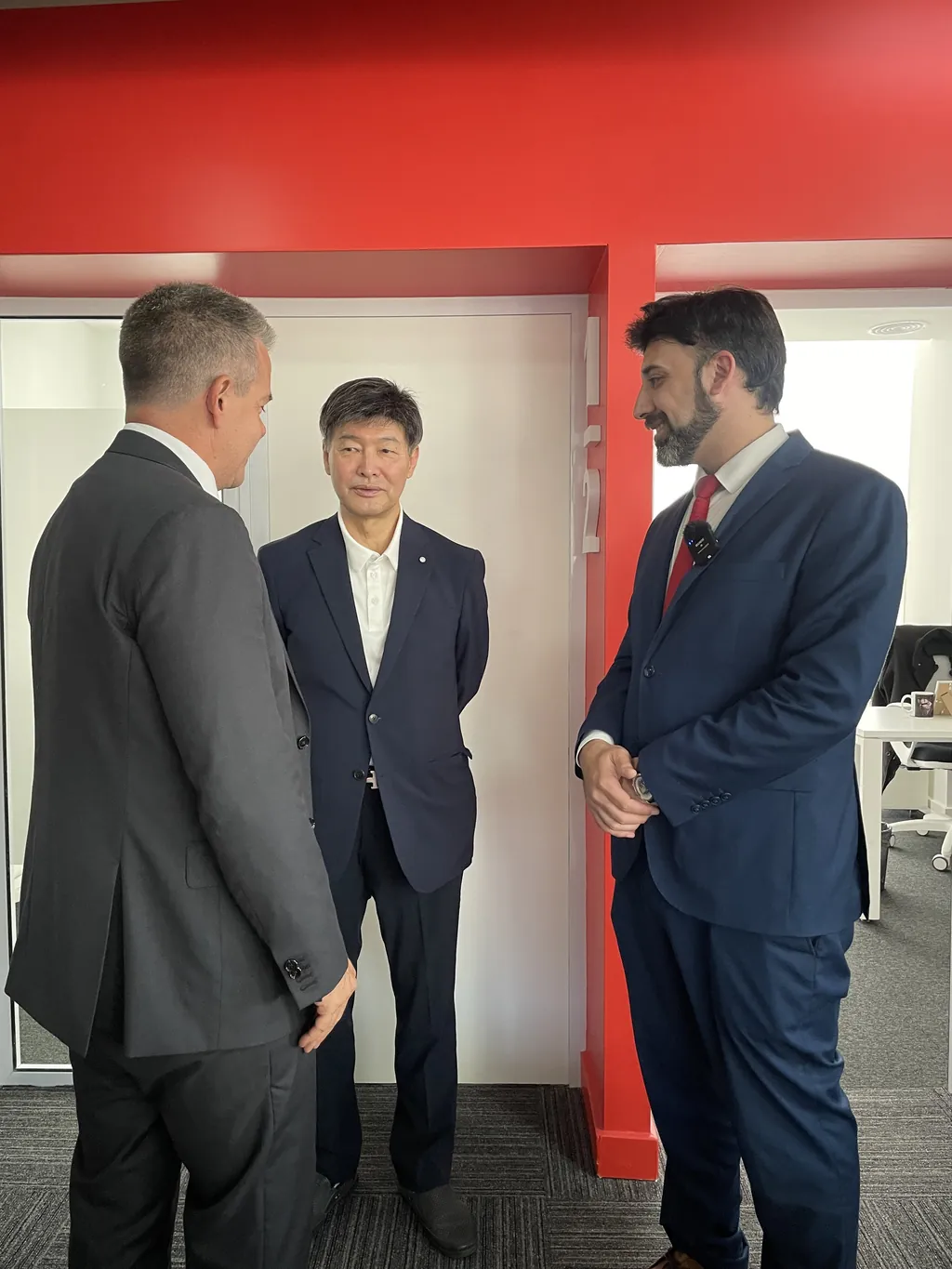 Tres personas intercambian ideas de pie, junto a una sala con paredes de vidrio.